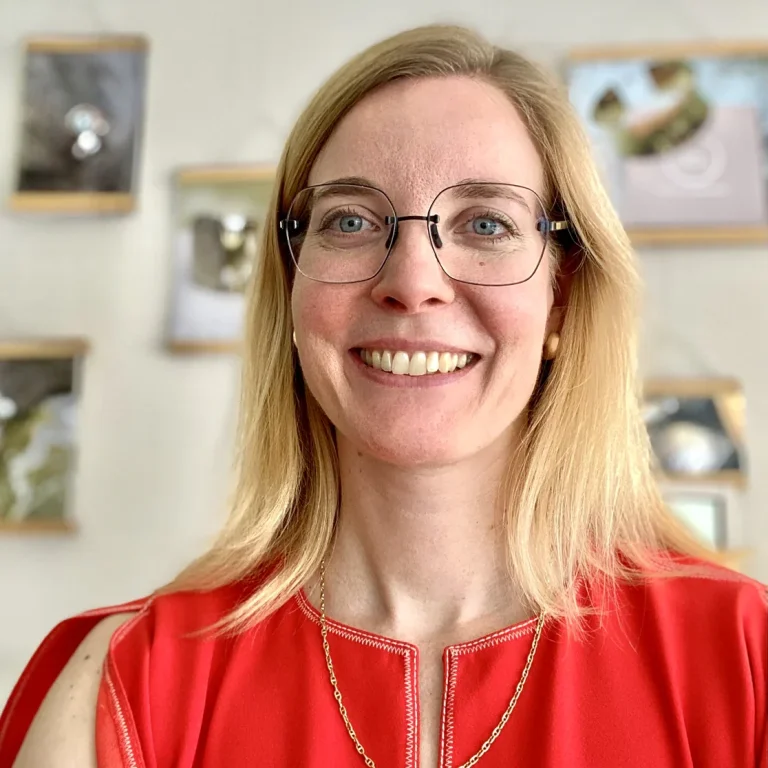 Lächelnde Frau mit blonden Haaren, rotem Oberteil und Brille vor Fotos im Hintergrund.