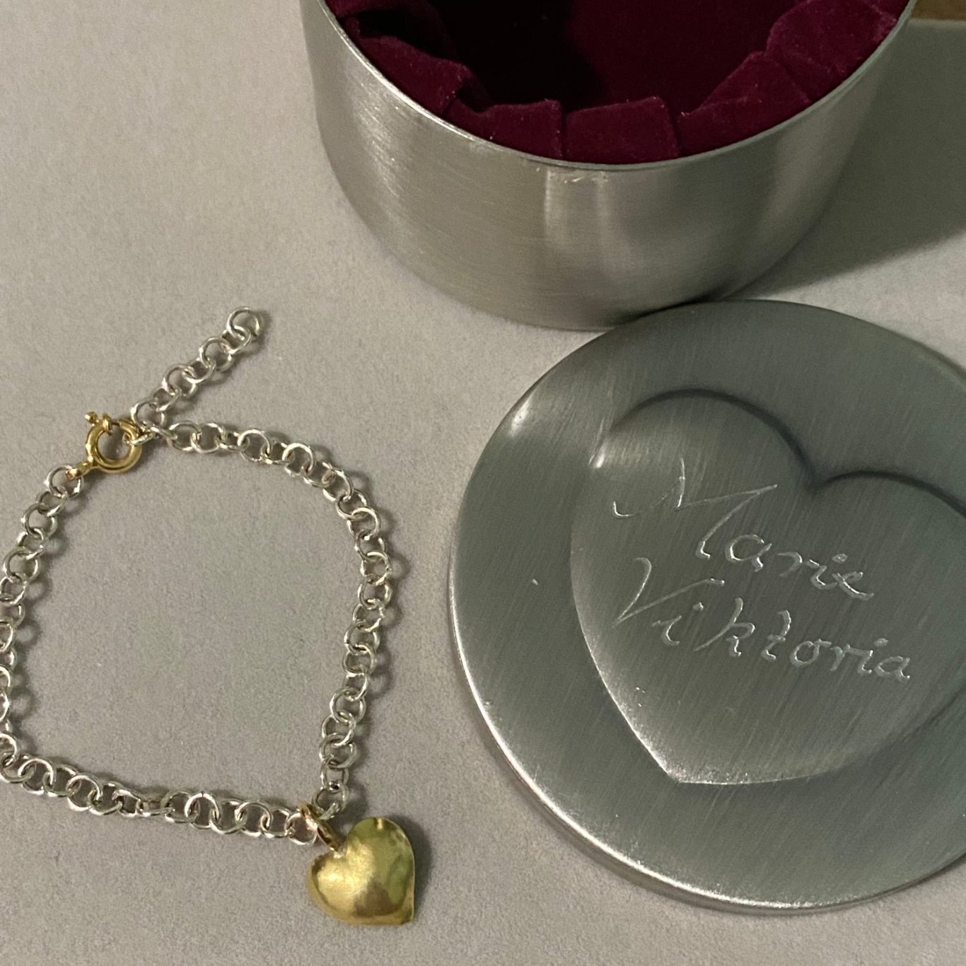 Silbernes Armband mit goldfarbenem Herzanhänger, neben einer runden Schmuckbox.