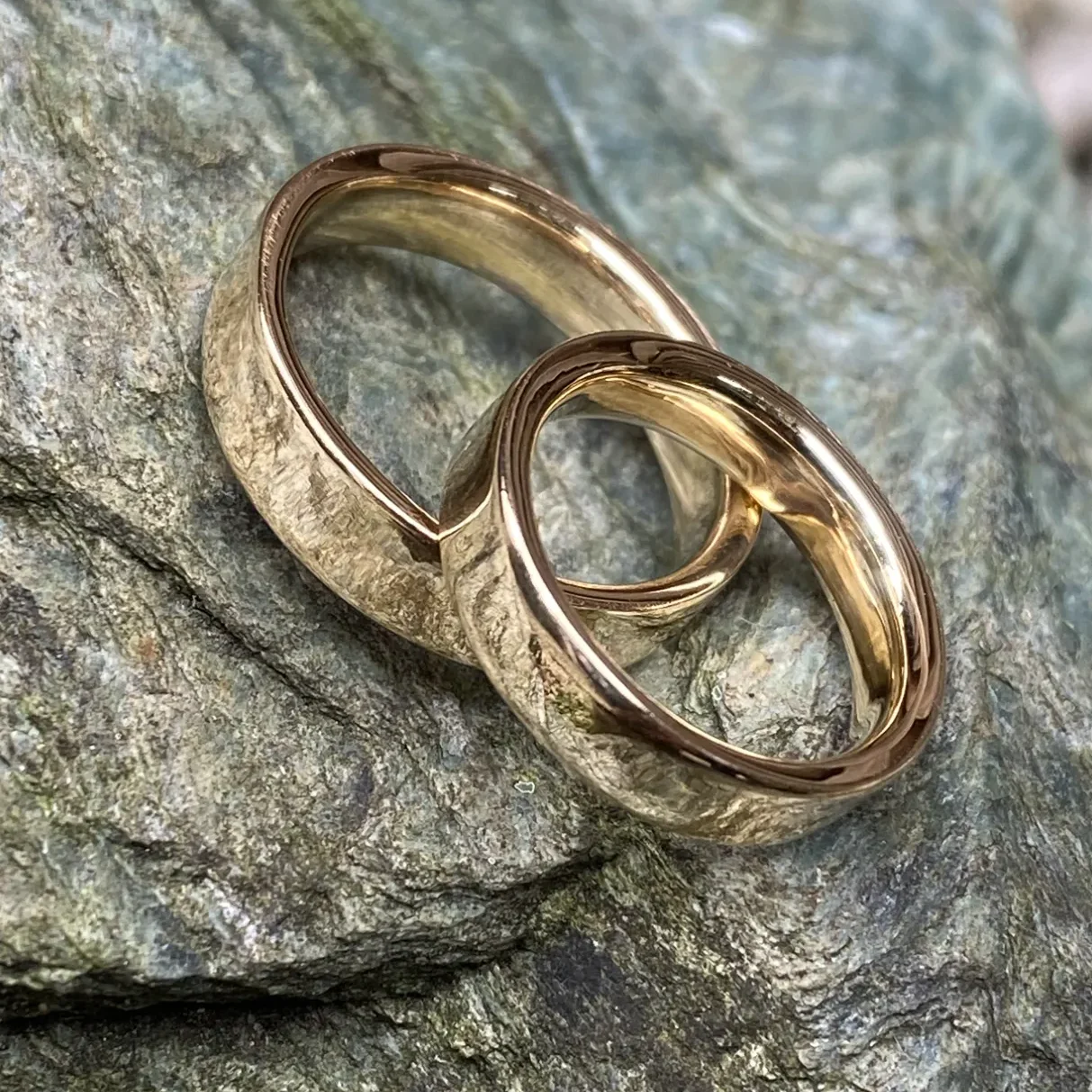 Zwei glänzende, goldene Trauringe liegen nebeneinander auf steinigem Untergrund.
