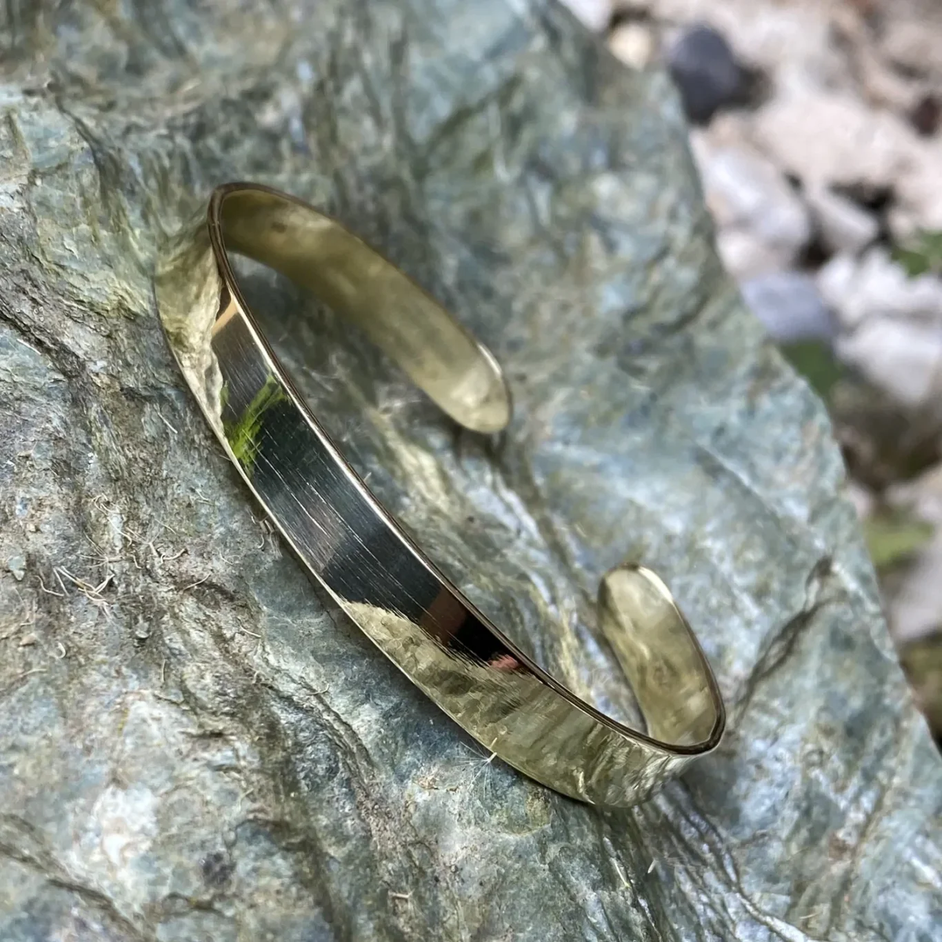 Goldfarbene schmale Armspange liegt auf einem steinigen Untergrund.