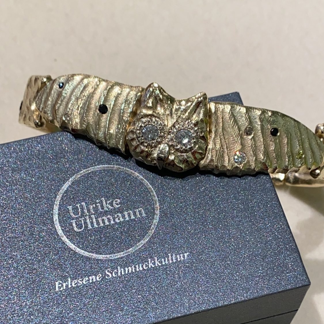 Goldenes Armband in Form einer Eule auf einer grauen Verpackung.