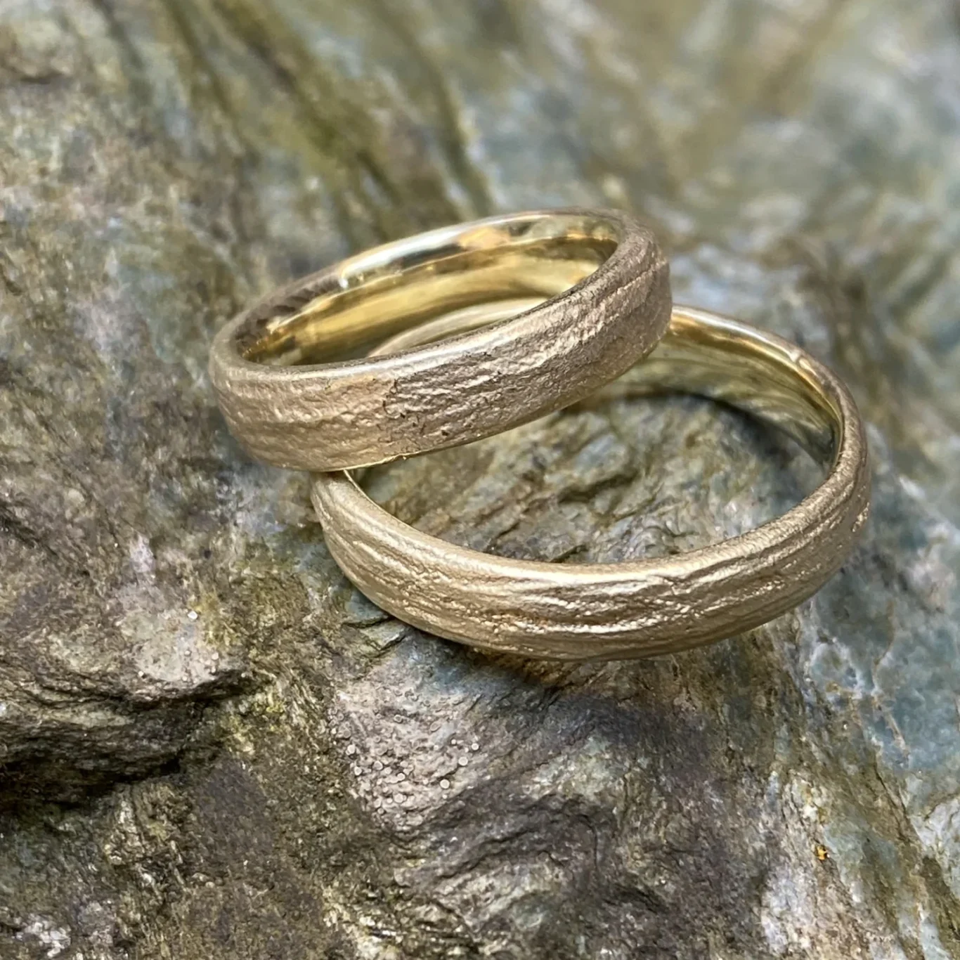 Zwei goldene Trauringe mit Holzstruktur auf einem Stein liegend.