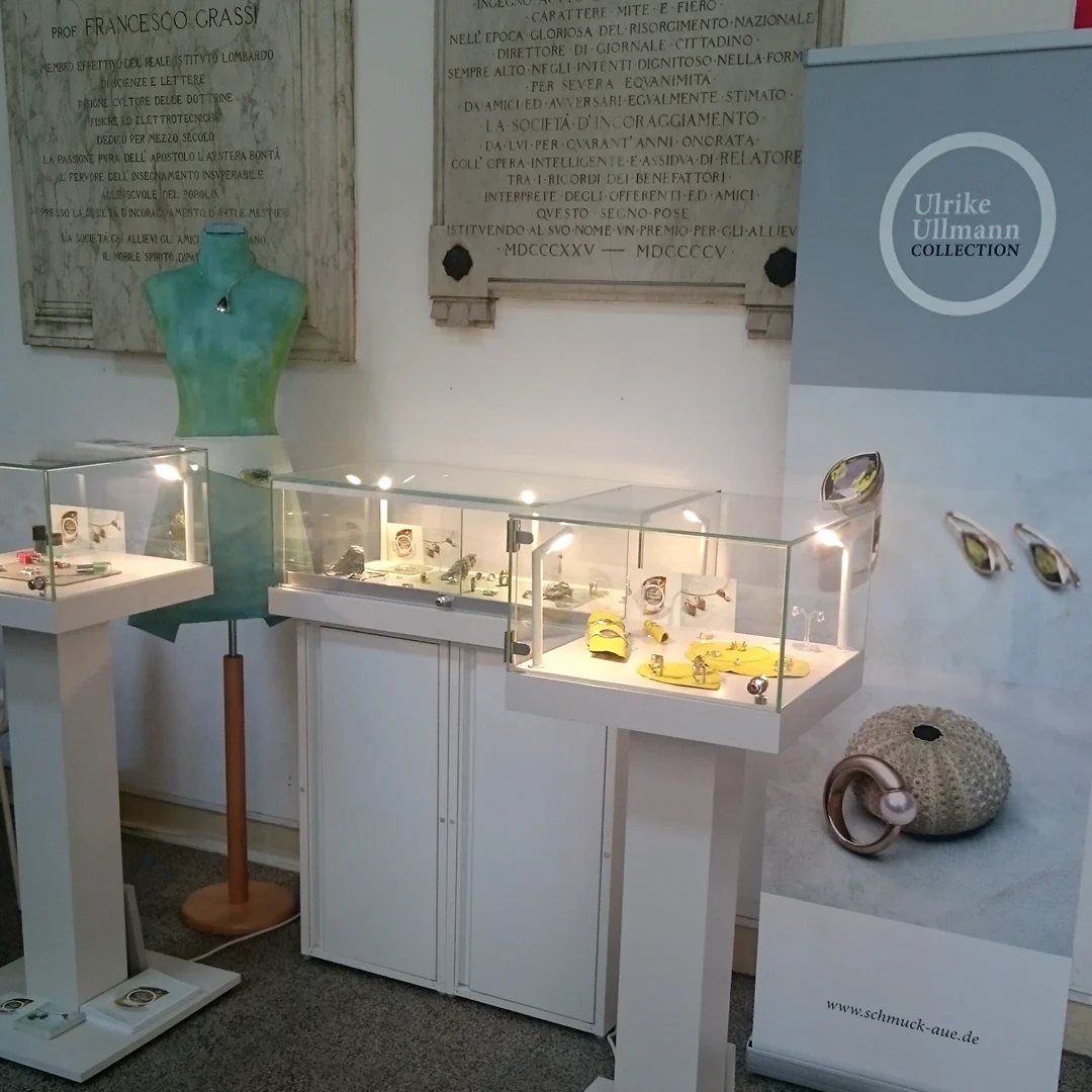 Milan Design Week Schmuckausstellung mit Vitrinen, einer Kleiderpuppe und einem Werbebanner im Hintergrund.