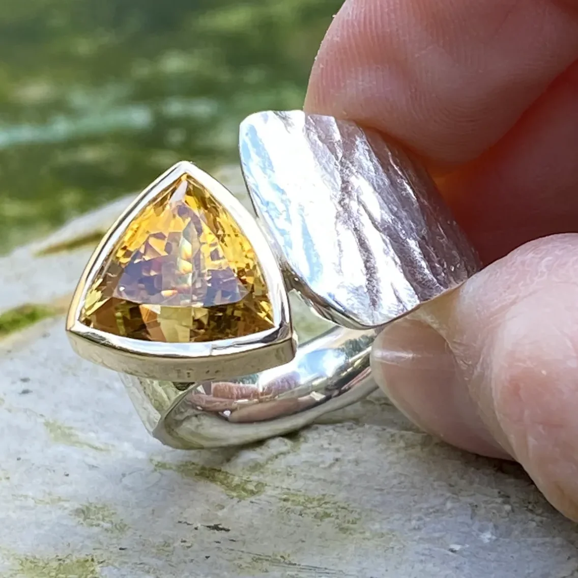 Ring aus Silber mit einem glitzernden gelben Citrin in einer Goldfassung mit zwei Fingern ins Licht gehalten, im Hintergrund ein Felsen.